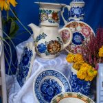 Zestaw porcelany z kwiatowym motywem na niebieskim tle. Dominują kolory: biel, niebieski, brązowy i różowy. Obok wazon z żółtymi kwiatami.