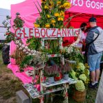 Stoisko z kwiatami i czosnkiem na tle różowego namiotu. Duże drewniane napisy Głęboczanki. Obok ludzie rozmawiający.