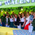 Grupa ludzi pod zielonym namiotem z napisem, ubrana w regionalne stroje. Tło ozdobione kwiatami. Na pierwszym planie żółte stoły.