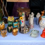 Stoisko z ceramiką: kolorowe sowy, anioły, dzbanki i dzwonki na białym obrusie. Tło ciemne, podkreślające barwy.