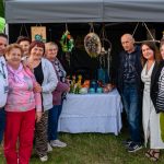 Grupa ludzi stoi przed namiotem z rękodziełem. Tło to zielony i szary namiot z kolorowymi dekoracjami i ceramiką. Wszyscy się uśmiechają.