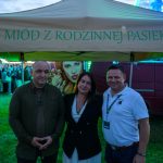 Trzy osoby stoją pod beżowym namiotem z napisem Miód z rodzinnej pasieki. W tle zielone i niebieskie światła oraz czerwony van. Trawa na pierwszym planie, a za osobami tłum ludzi.