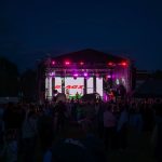 Nocny koncert na scenie z fioletowymi światłami. Tłum widzów z przodu, drzewa w tle. Wyświetlacz z napisem na scenie.
