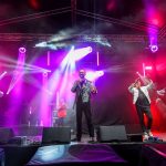 Trzech wykonawców na scenie w kolorowym oświetleniu. Jasne reflektory i fioletowe światła tworzą dynamiczną atmosferę. Metalowa konstrukcja nad nimi podkreśla koncertowy klimat.