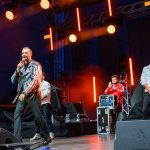 Mężczyzna w kolorowej kurtce śpiewa na scenie. Tło to wieczorne oświetlenie z pomarańczowymi reflektorami. Obok niego stoją dwaj inni mężczyźni.