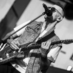 Muzyk w okularach i kamizelce gra na gitarze elektrycznej, śpiewając do mikrofonu. Czarnobiałe zdjęcie na scenie. Tło rozmyte, jasne.