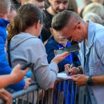 Mężczyzna w okularach podpisuje autograf przy barierce. Obok niego stoi osoba w szarej bluzie, trzymająca telefon. Tłum w tle, dominujące kolory to niebieski i szary.