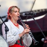 Kobieta w białej bluzce i czerwonej spódnicy gra na klarnecie na scenie. Tło jest ciemne, z mikrofonem z przodu. Widać detale instrumentu.