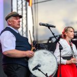 Dwóch muzyków występuje na scenie; mężczyzna w kapeluszu gra na bębnie, kobieta w czerwonej spódnicy gra na klarnecie. Scena jest na zewnątrz, pod niebieskim niebem. Mikrofony stojące przed nimi podkreślają atmosferę koncertową.