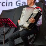 Mężczyzna w kapeluszu gra na akordeonie, siedząc na stołku. Tło jest czarne, z widocznymi napisami. Stojak na nuty po lewej.