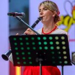 Osoba gra na klarnecie, ubrana w białą bluzkę i czerwoną spódnicę. Tło jest kolorowe, z mikrofonem obok. Przed nią stoi pulpit nutowy.