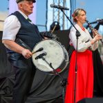 Dwóch muzyków na scenie, mężczyzna gra na bębnie, a kobieta na klarnet. Kobieta ma czerwoną spódnicę, mężczyzna jasną koszulę z kamizelką. Tło zewnętrzne, jasne niebo i scena oświetlona światłami.