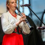 Kobieta w stroju ludowym gra na klarnecie, ubrana w białą bluzkę i czerwoną spódnicę. W tle sceniczne oświetlenie. Instrument zbliżony do mikrofonu.