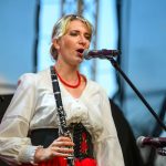 Kobieta w stroju ludowym gra na klarnecie na scenie. Tło niebieskie, mikrofon przed nią. Noszą czerwoną spódnicę i naszyjnik.