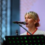 Kobieta mówi do mikrofonu na scenie. Ubrana w białą bluzkę i czerwone korale. Tło jest rozmyte i jasnoszare.