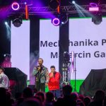 Na scenie trzech mężczyzn, jeden śpiewa. W tle jasne ekrany z tekstem i kolorowe reflektory. Na widowni ciemne sylwetki.