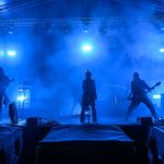 Na scenie w niebieskim świetle występuje zespół muzyczny. Przód zajmuje wokalista, otoczony przez gitarzystów. Tło wypełnia gęsty dym sceniczny.