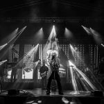 Na zdjęciu jest zespół rockowy na scenie. Dominuje czarno-biały kontrast i intensywne reflektory. W tle widać ekran z dużym wizerunkiem osoby.