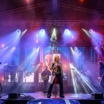 Koncert na scenie z zespołem, dymem i kolorowymi reflektorami w tle. Przeważają kolory fioletowy i niebieski. Wokalistka stoi na środku z mikrofonem.