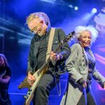 Na scenie para muzyków gra na gitarach. Tło jest fioletowe z jasnymi światłami. Artyści noszą ciemne ubrania i wykazują energię.