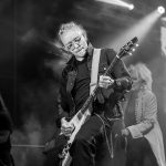 Mężczyzna gra na gitarze elektrycznej na scenie, oświetlony reflektorami. Tło jest ciemne z efektami dymu. Zdjęcie wykonane w czerni i bieli.