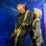 Mężczyzna gra na gitarze na scenie, ubrany na czarno. Obok niego kobieta w szarym płaszczu z białymi włosami. Tło jest ciemne z niebieskimi światłami.