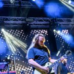 Muzyk gra na gitarze elektrycznej na scenie. Oświetlenie w tle jest kolorowe i dynamiczne. Scena jest pełna reflektorów i dymu.