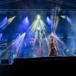 Zespół rockowy na scenie, kolorowe światła i dymy. Centralna postać śpiewa, otoczona gitarzystami. Tło jest ciemne.
