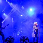 Koncert rockowy z gitarzystą i wokalistką na scenie. Dominują niebieskie światła i dym, tworzące dynamiczną atmosferę. W tle perkusja i intensywne oświetlenie.