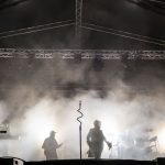 Koncert rockowy na scenie, muzycy w dymie podświetlonym białym światłem, metalowa konstrukcja w tle.