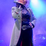Osoba na scenie z mikrofonem, w jasnym płaszczu, na tle fioletowych świateł. Ma białe włosy i bogato zdobiony pasek.