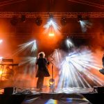 Koncert na scenie z muzykami wśród kolorowych świateł. Dominują pomarańczowe i białe reflektory. Dym na tle dodaje dynamiki.