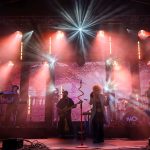 Koncert zespołu na scenie oświetlonej czerwonymi i białymi reflektorami. Dym unosi się w tle, tworząc dynamiczną atmosferę. Muzycy grają na gitarach i instrumentach elektrycznych.