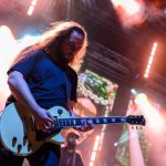 Muzyk z gitarą elektryczną na scenie, otoczony kolorowymi światłami. W tle ekran i aparatura. Scena w dynamicznych barwach.