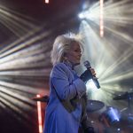 Osoba śpiewa na scenie trzymając mikrofon. Tło ma efektowne światła w różnych kolorach. Widać bębny perkusyjne.