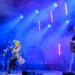 Na scenie artysta z kwiatowym wieńcem śpiewa. Neonowe światła w odcieniach niebieskich i fioletowych tworzą efekt tła. Muzycy grają na gitarach przy jego boku.