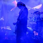 Muzyk gra na gitarze na scenie w niebieskim świetle, obok perkusista i klawiszowiec. Dym i efekty świetlne w tle.