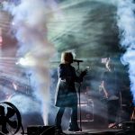 Na scenie widoczny jest wokalista z mikrofonem i gitarzysta. Dym i kolorowe światła tworzą dynamiczny klimat koncertu. Tło mieni się pastelowymi odcieniami.