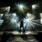 Koncert rockowy na scenie z intensywnym oświetleniem. Światła w różnych kolorach tworzą dynamiczny efekt. Muzycy są w cieniu, a tło jest wypełnione mgłą.