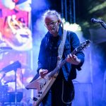 Muzyk gra na gitarze na scenie podczas koncertu. Tło jest kolorowe z intensywnym oświetleniem. Widać instrumenty wśród mgły.