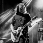Mężczyzna gra na gitarze elektrycznej na scenie, oświetlony jasnym światłem reflektorów. Tło to dym i sprzęt muzyczny. Zdjęcie jest czarno-białe.