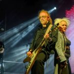 Na zdjęciu duet muzyczny na scenie: gitarzysta i wokalistka. Jasne reflektory oświetlają scenę, a tło jest ciemne. Kolorystyka ciepła, akcenty czerwienia.