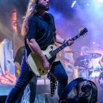 Mężczyzna z długimi włosami gra na kremowej gitarze na scenie. Tło rozświetlone efektami świetlnymi w odcieniach niebieskiego i fioletu. Przed nim znajduje się wentylator, a w tle widać perkusistę.
