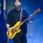Muzyk gra na żółtej gitarze basowej na scenie. Nosi czapkę z daszkiem i ciemną koszulę. Tło jest niebieskie, sceniczne.