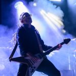 Muzyk gra na gitarze elektrycznej w jasnym świetle reflektorów i mgiełce. Kolory dominujące to niebieski i fioletowy. Tło jest zamglone, ozdobione światełkami.