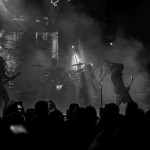 Na czarno-białym zdjęciu widzimy zespół rockowy na scenie w otoczeniu dymu. Muzycy z gitarami i perkusją, intensywne światła w tle. Publiczność z przodu w cieniu.