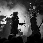 Na czarno-białym zdjęciu zespół rockowy na scenie. Wokalista z mikrofonem i basista z gitarą w dymie. Tło jest ciemne i rozmyte.