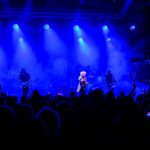 Muzyka na żywo na scenie, oświetlenie w intensywnym niebieskim kolorze, publiczność w ciemnościach na pierwszym planie.