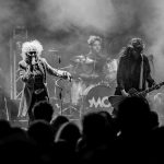 Na czarno-białym zdjęciu grupa rockowa występuje na scenie. Wokalista trzyma mikrofon, gitarzysta z gitarą. W tle perkusista i publiczność.