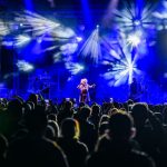 Koncert na żywo na scenie z niebieskimi światłami i dymem. Artysta w centrum, widownia z telefonami w górze. Tło w dynamice świateł.
